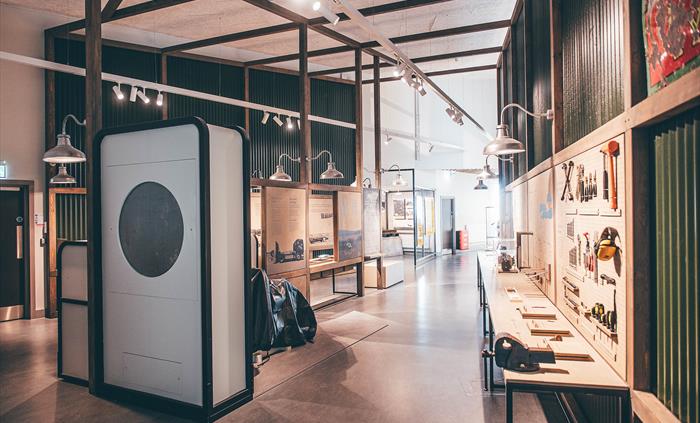 A view of a gallery space with a workbench and tools on the right and wind tunnel apparatus on the left.